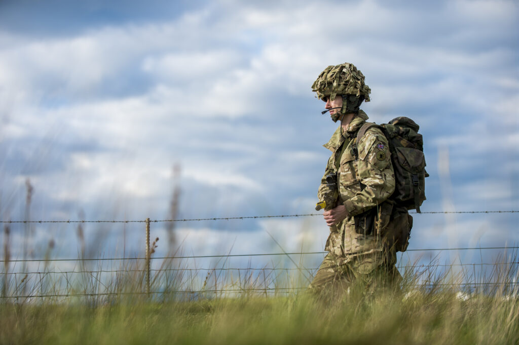 A soldier is photographed against a blue sky, walking on patrol. He is in uniform, wearing a bergen and helmet and carrying a rifle.