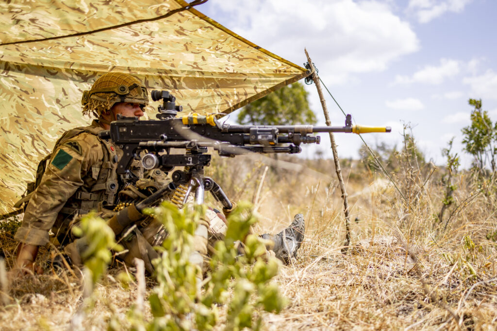 A soldier sits in dry grassland, aiming a machine gun. He is covered by a camouflage tarp rigged above his head for shade.