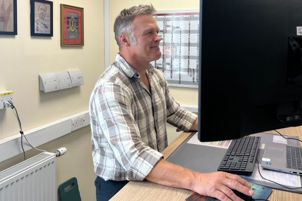 A man at a standing desk with a computer. He has one hand on the mouse and is looking at the monitor. He wears civilian clothes.