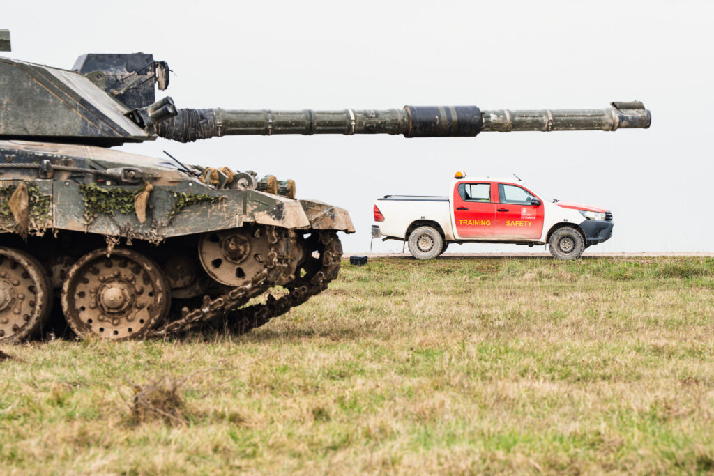 On the left of the image is the front of a tank on grass. The tank's barrel points horizontally across the image, and just underneath this but much further back is a red and white pickup truck marked 'Training Safety'.